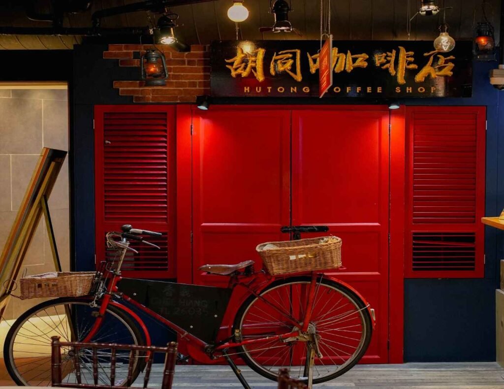 The old red bicycle with baskets, parked in front of a rustic Asian wooden shopfront door and window with red lanterns and a vintage Chinese signboard in Lot 10 Hutong Food Court. Visitors enjoy snapping Instaworthy photos in this charming old-school setting.