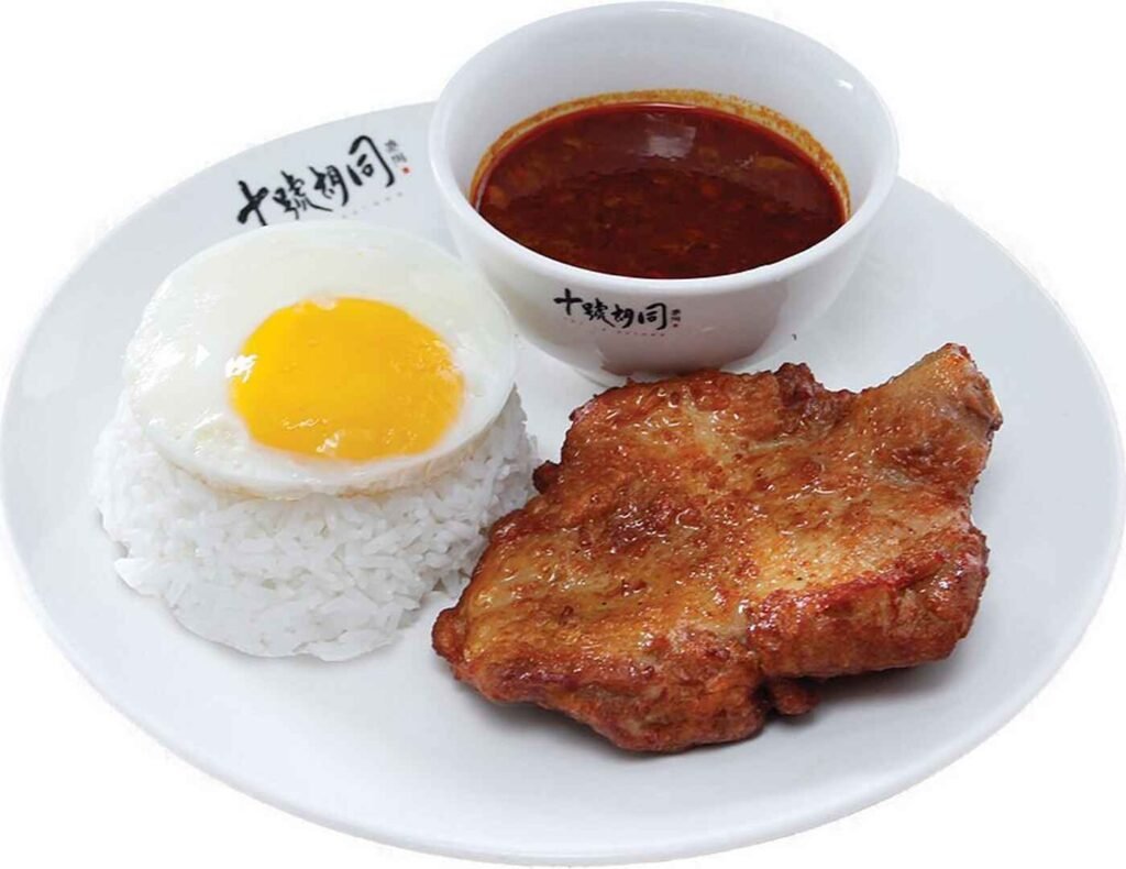 The Roasted Pork Rice With Egg features crispy-skinned roasted pork, fragrant white rice, topped by a sunny side up egg and a side of spicy-savoury curry, offering a comforting taste of classic Hong Kong-style hawker fare. A signature dish at Loong Stall, Lot 10 Hutong.
