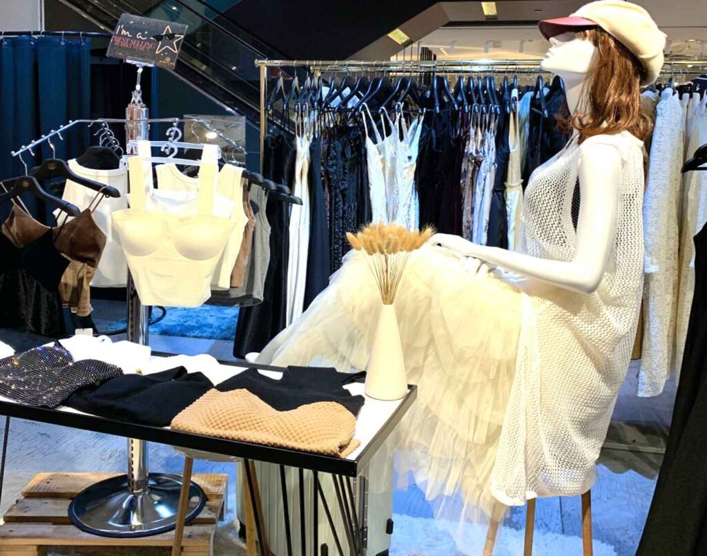 Bubbly You display featuring a mannequin wearing a white mesh sleeveless tunic over a white bra top and a sheer, pleated tulle skirt, embodying the K-fashion aesthetic. On a table nearby, black and beige knit sweaters are neatly folded next to a gold-embellished baseball cap and a white vase holding dried grass. The surrounding clothing racks hold a variety of casual wear, including camisoles, shorts, and trousers. Located at The Fashion, First Floor, Isetan The Japan Store Lot 10, Bukit Bintang, Kuala Lumpur.