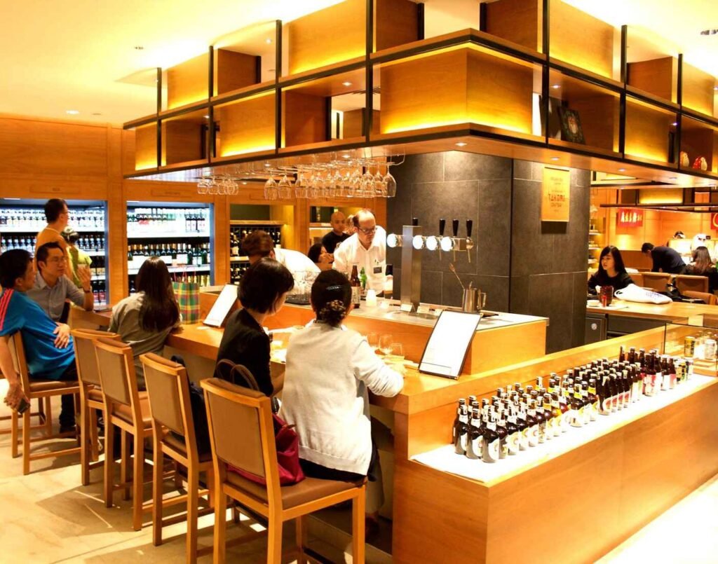 One of the drinking bars of City Cellar, a well-lit wooden bar with patrons seated on stools and staff serving them. The bar features a display of wine and liquor bottles and beverage taps. The background includes a large refrigerated display and a modern, cube-shaped shelving unit filled with imported alcoholic beverages. The warm lighting, elegant ambience and the variety of wines, spirits, liquors and craft beers at City Cellar, located at The Market, Lower Ground Floor, Isetan The Japan Store Lot 10, Bukit Bintang, Kuala Lumpur, makes it a favourite drinking spot for after-work professionals and tourists alike.