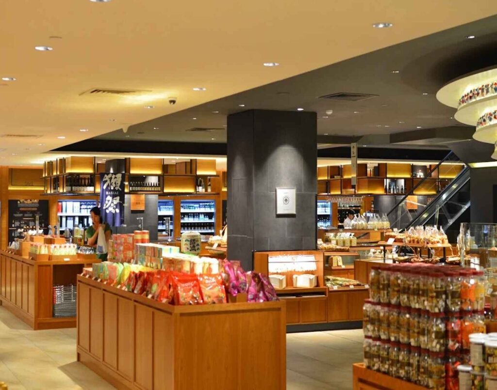 The Market features fresh groceries, artisanal Japanese snacks and specialty items. Bathed in warm, clean lighting, the polished wooden display stands create a premium shopping atmosphere, while the shelves showcase an impressive variety of products — from colorful Japanese snacks and sweets in the foreground to an extensive selection of sake, wine, and bottled beverages neatly arranged in the background. Located on the Lower Ground Floor, Isetan The Japan Store Lot 10, Bukit Bintang, Kuala Lumpur.