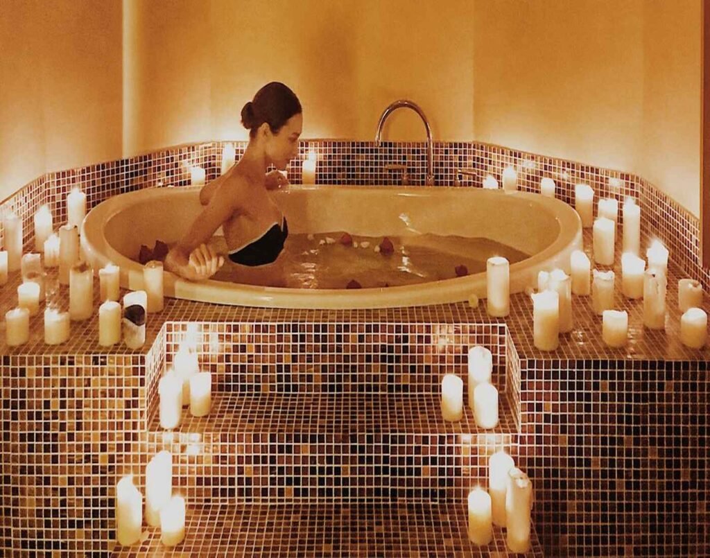 A woman relaxing in a warm candlelit bath surrounded by rose petals and dozens of glowing candles, under an ornate Moroccan archway with a brass lantern casting soft patterns on the walls. This relaxing hot bath is part of the My Favourite Concubine package, available at Hammam Spa, The Wellness, Second Floor, Isetan The Japan Store Lot 10, Bukit Bintang, Kuala Lumpur.