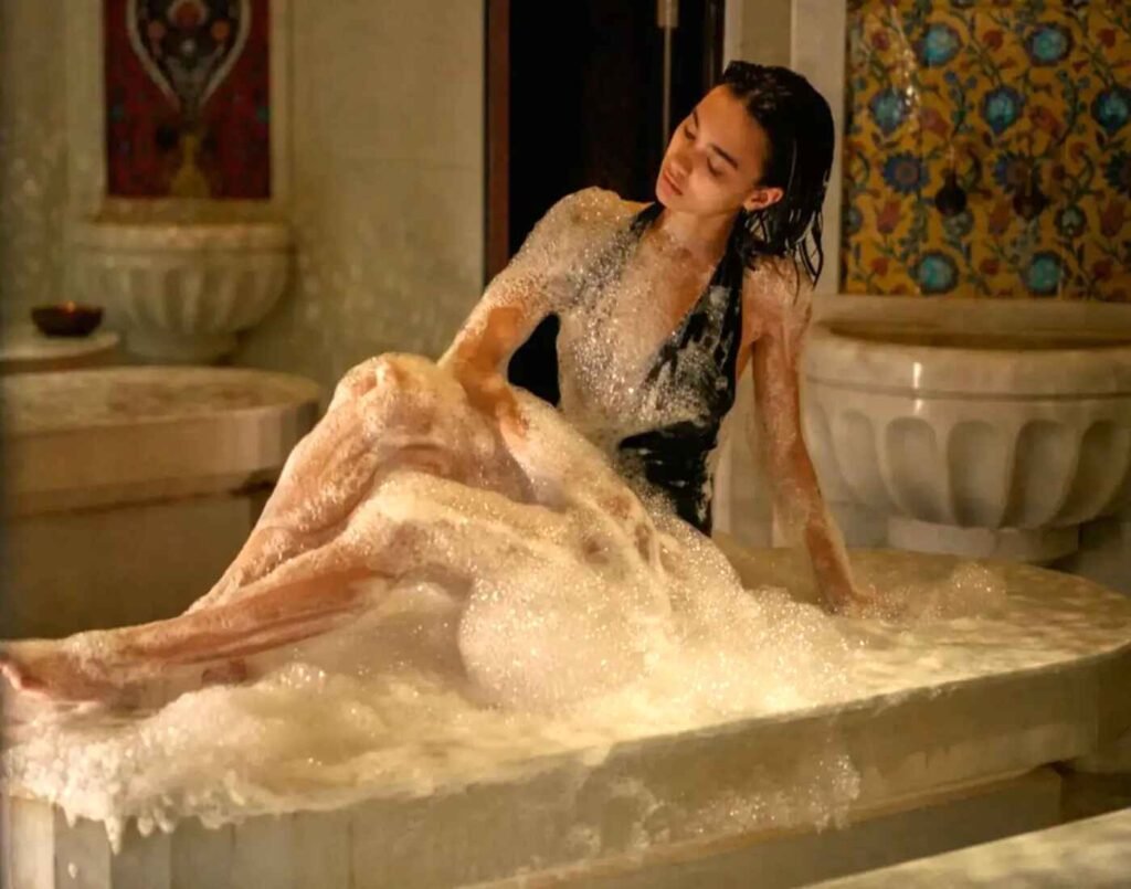 A woman enjoying the traditional Moroccan foam bath ritual at Hammam Spa, covered in bubbles while seated on a heated Carrara marble bed, with mosaic wall tiles and carved stone basins in the background. Located at The Wellness, Second Floor, Isetan The Japan Store Lot 10, Bukit Bintang, Kuala Lumpur.