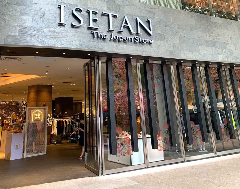 Exterior view of Isetan The Japan Store’s main entrance with a large backlit ‘ISETAN The Japan Store’ sign on a stone facade, full-height glass doors, and floral window panels. Inside, mannequins and pop-up displays create a warm, welcoming storefront. Located in Lot 10 Mall, Bukit Bintang, Kuala Lumpur.