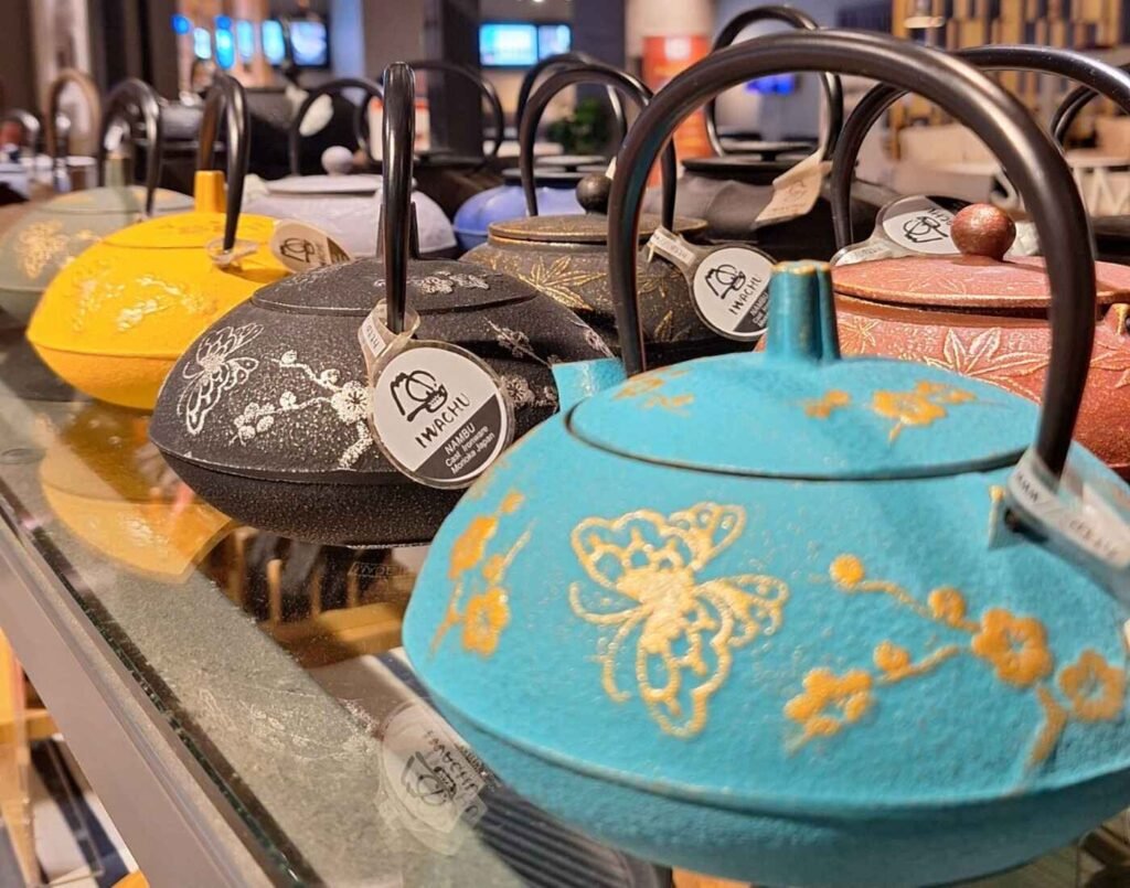 A row of colourful Iwachu Japanese cast-iron teapots (tetsubin) with intricate floral and butterfly motifs, showcasing traditional Nambu Ironware craftsmanship. Displayed in vibrant shades of yellow, black, and turquoise at The Design, Ground Floor, Isetan The Japan Store Lot 10, Bukit Bintang, Kuala Lumpur.