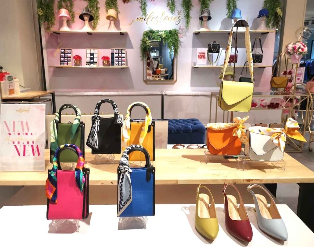 Milestones365 boutique display showcasing structured mini handbags in bold shades of pink, blue, green, yellow, orange, white, and black, each adorned with patterned scarf handles. Below, pointed-toe slingback shoes in muted tones of mustard, maroon, and dove grey complete the look, while pastel hats and additional accessories line the background shelves. The setup reflects Milestones365’s playful yet polished approach to handbags, footwear, and fashion accents at The Fashion, First Floor, Isetan The Japan Store Lot 10, Bukit Bintang, Kuala Lumpur.
