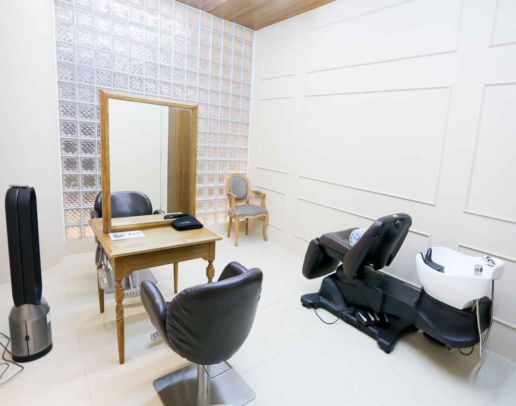 Number 76 salon interior, featuring a clean, minimalist aesthetic with a black leather treatment chair and a separate hair-washing station. The room is brightly lit, with white-paneled walls and a unique feature wall made of textured glass blocks, highlighting the modern and professional ambiance of the salon, located at The Wellness, Second Floor, Isetan The Japan Store Lot 10, Bukit Bintang, Kuala Lumpur.