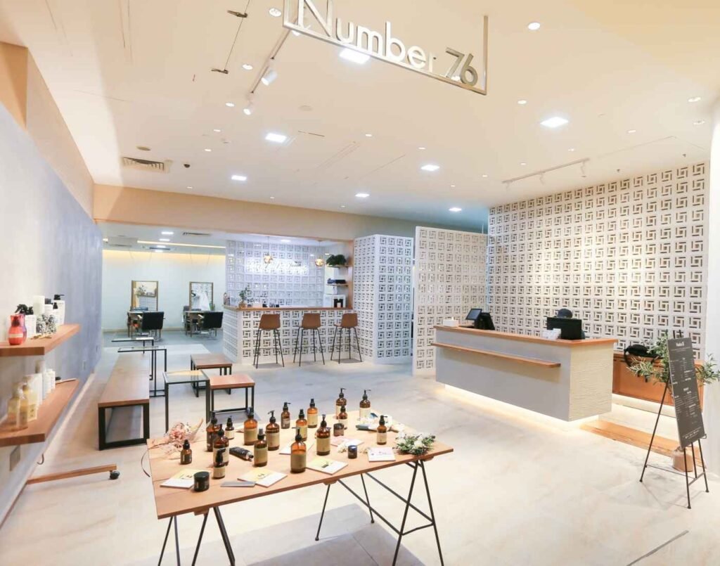 The reception and product display area of Number 76 salon. A large wooden table showcases an array of hair care products in amber bottles, while a clean white counter serves as the reception desk. The brand's name is visibly displayed in sleek, modern lettering, and a white, textured block wall adds a unique design element to the bright and inviting space. Located at The Wellness, Second Floor, Isetan The Japan Store Lot 10, Bukit Bintang, Kuala Lumpur.