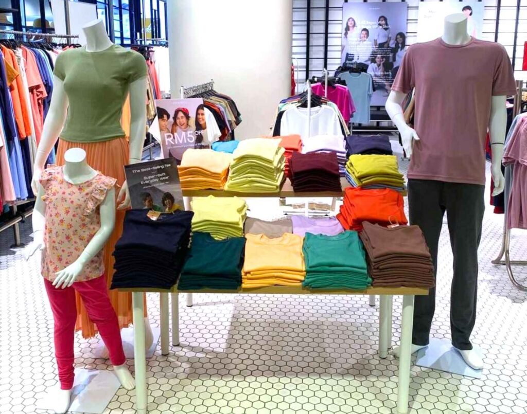 Oxwhite display featuring men’s, women’s, and children’s mannequins dressed in casual essentials, surrounded by stacks of neatly folded crew-neck T-shirts in a full spectrum of colours including navy, yellow, green, pink, orange, and beige. The presentation highlights Oxwhite’s focus on versatile, affordable, and comfortable everyday basics for the whole family at The Fashion, First Floor, Isetan The Japan Store Lot 10, Bukit Bintang, Kuala Lumpur.
