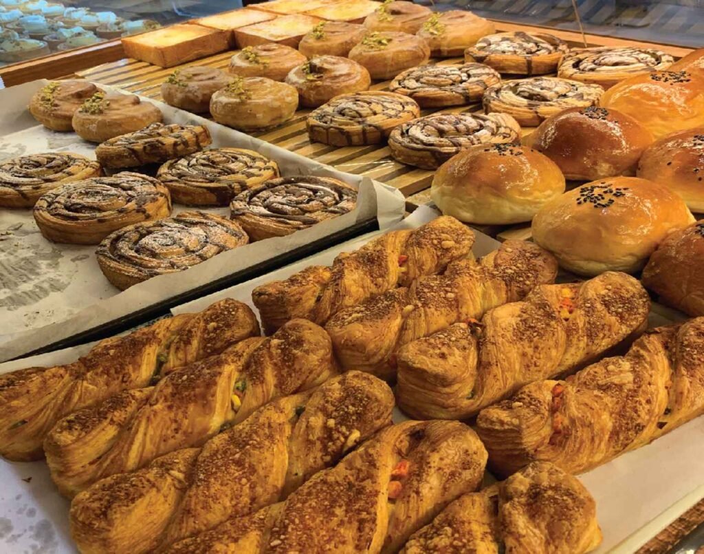 A shelf at Sol Levante Bakery displaying Braided Cheese Twists – flaky golden pastries with a light crumb topping and hints of savoury filling, Cinnamon Rolls – soft, spiral-shaped buns dusted with powdered sugar for a sweet finish, Chocolate Swirl Danish – rich, layered pastries drizzled or filled with chocolate, Glazed Milk Buns – fluffy, golden rolls, some topped with black sesame seeds for extra flavour, Pistachio Danish – glossy, round pastries finished with a sprinkle of chopped pistachios, Japanese Milk Bread (Shokupan) – square-shaped, soft loaves with a tender crumb, perfect for toasting; located at The Market, Lower Ground Floor, Isetan The Japan Store Lot 10, Bukit Bintang, Kuala Lumpur.