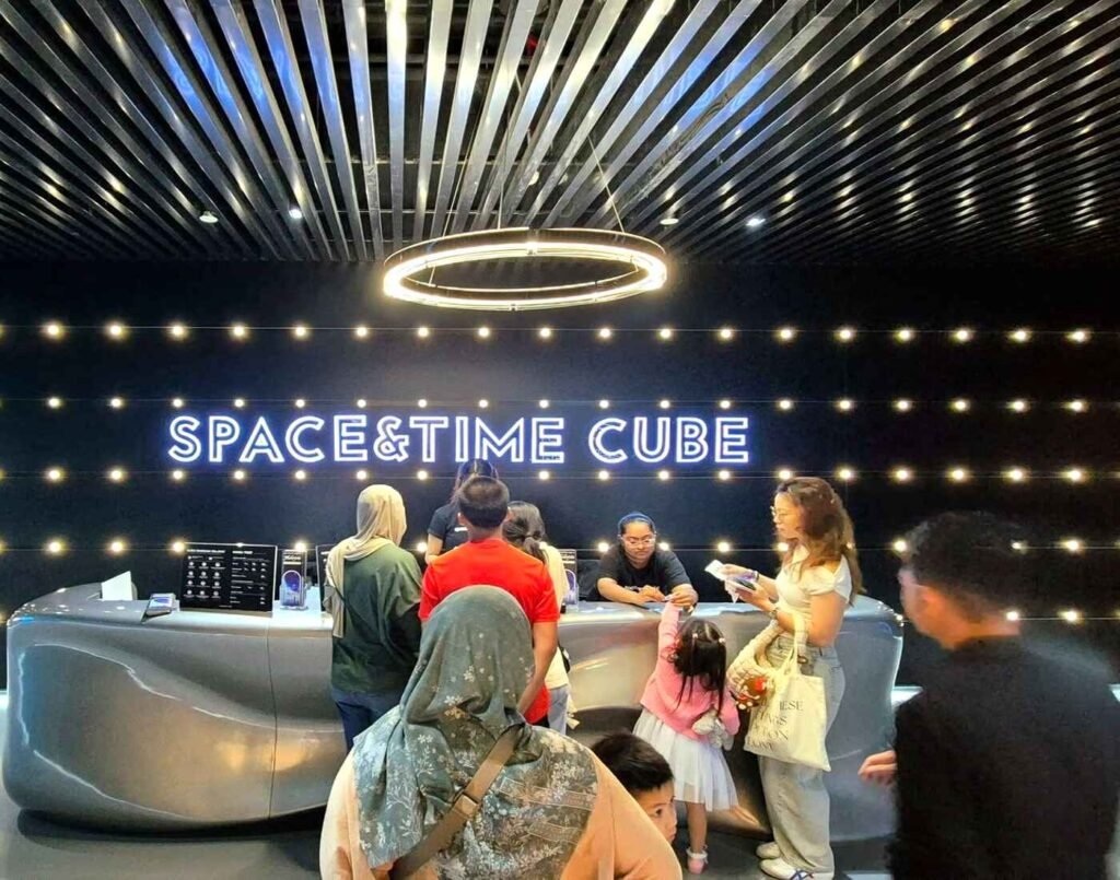A modern, dark-themed reception and ticketing counter at the entrance of the Space & Time Cube. The wall behind the sleek, curved grey counter features a prominent, backlit logo that reads "SPACE & TIME CUBE" flanked by a grid of glowing, circular lights. Several customers, including families, are queueing at the counter to purchase tickets from the staff. Located at The Amusement, Third Floor, Isetan The Japan Store Lot 10, Bukit Bintang, Kuala Lumpur.