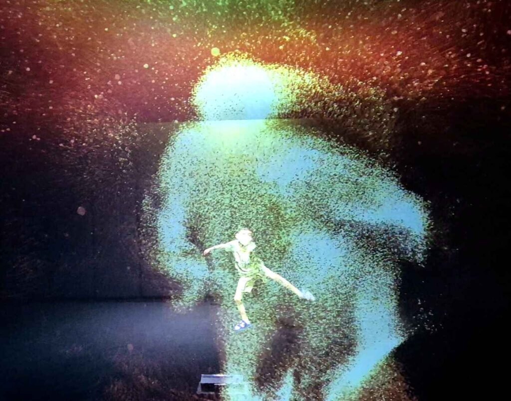 A child jumps and moves in front of a large screen in a dark room. The screen displays a shimmering, particle-based silhouette of the child that moves in real time, creating a glowing yellow-green coloured Doppelganger effect. This interactive shadow projection is part of the immersive experience at Space & Time Cube, The Amusement, Third Floor, Isetan The Japan Store Lot 10, Bukit Bintang, Kuala Lumpur.