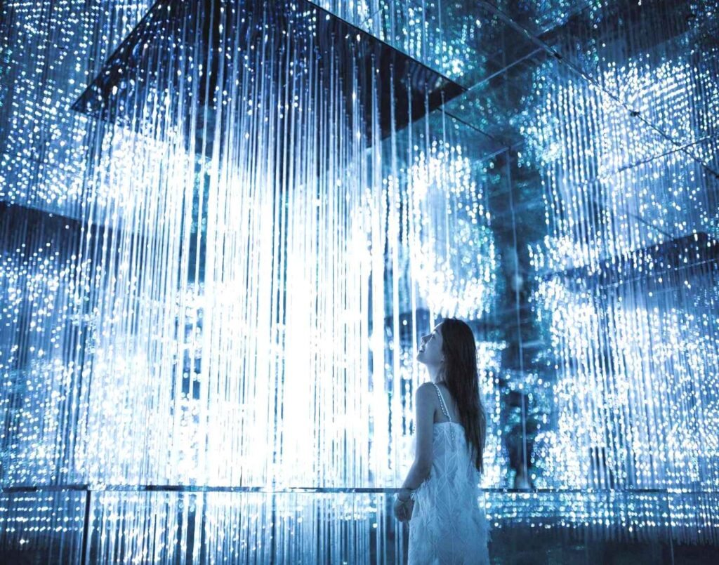 A woman looking up at thousands of luminous crystal-shaped LED light strips suspended from the ceiling, resembling a celestial shower of radiant stars at the Space Puzzle. The lights are a cool, glowing blue and white. The entire zone, including the floor and walls, is mirrored, causing the light fixtures to appear to stretch into infinity in every direction. A highlight at Space & Time Cube, The Amusement, Third Floor, Isetan The Japan Store Lot 10, Bukit Bintang, Kuala Lumpur.