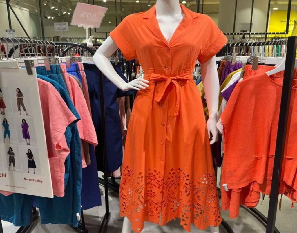 WhattoWear display set featuring a mannequin in a stylish, bright orange, short-sleeve button-down dress with a tie-waist and intricate laser-cut floral patterns on the skirt hem, surrounded by clothing racks with various colourful and versatile skirts, dresses, tops and women’s apparel, located at The Fashion, First Floor, Isetan The Japan Store Lot 10, Bukit Bintang, Kuala Lumpur.