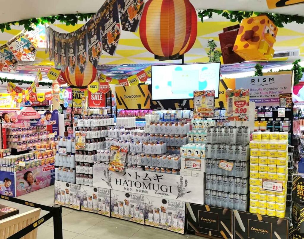 CosmeDONKI skincare displays featuring Naturie Hatomugi skin conditioner, RISM sheet masks, and MegRhythm steam eye masks, arranged in large promotional stacks adjacent to the dining area on the 2nd floor of Don Don Donki Lot 10, a Japanese supermarket in Bukit Bintang, Kuala Lumpur.