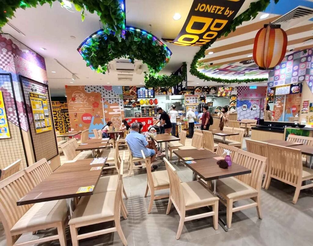 Wooden dining tables and chairs at the Don Don Donki Lot 10 dining area surrounded by Japanese specialty food stalls serving sushi, onigiri and donburi.