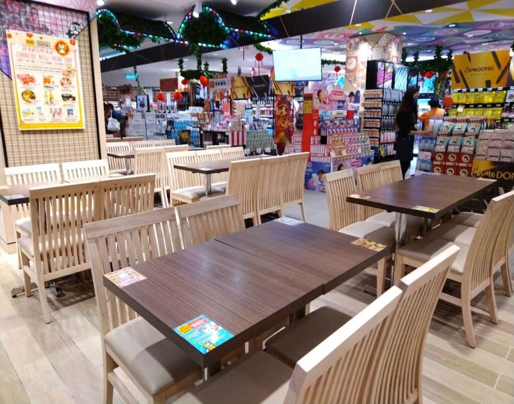 A dedicated seating area for customers of the various food vendors on the second floor. It features wooden tables and light wood banquette seating, providing a casual space to enjoy freshly prepared meals. Located on the second floor at Jonetz By Don Don Donki Lot 10, a Japanese supermarket in Bukit Bintang, Kuala Lumpur.