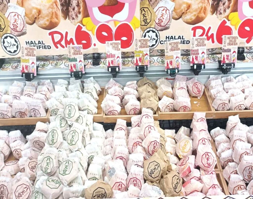 A refrigerated display case fully stocked with Halal-certified Hattendo Chilled Cream Buns in paper wrapping, with a large price tag reading RM6.90. Each palm-sized bun is filled with cold, creamy custard and available in five different flavours: Original Custard, Matcha, Azuki Beans, Chocolate, and Whipped Cream. Available at Jonetz By Don Don Donki Lot 10, a Japanese supermarket in Bukit Bintang, Kuala Lumpur.
