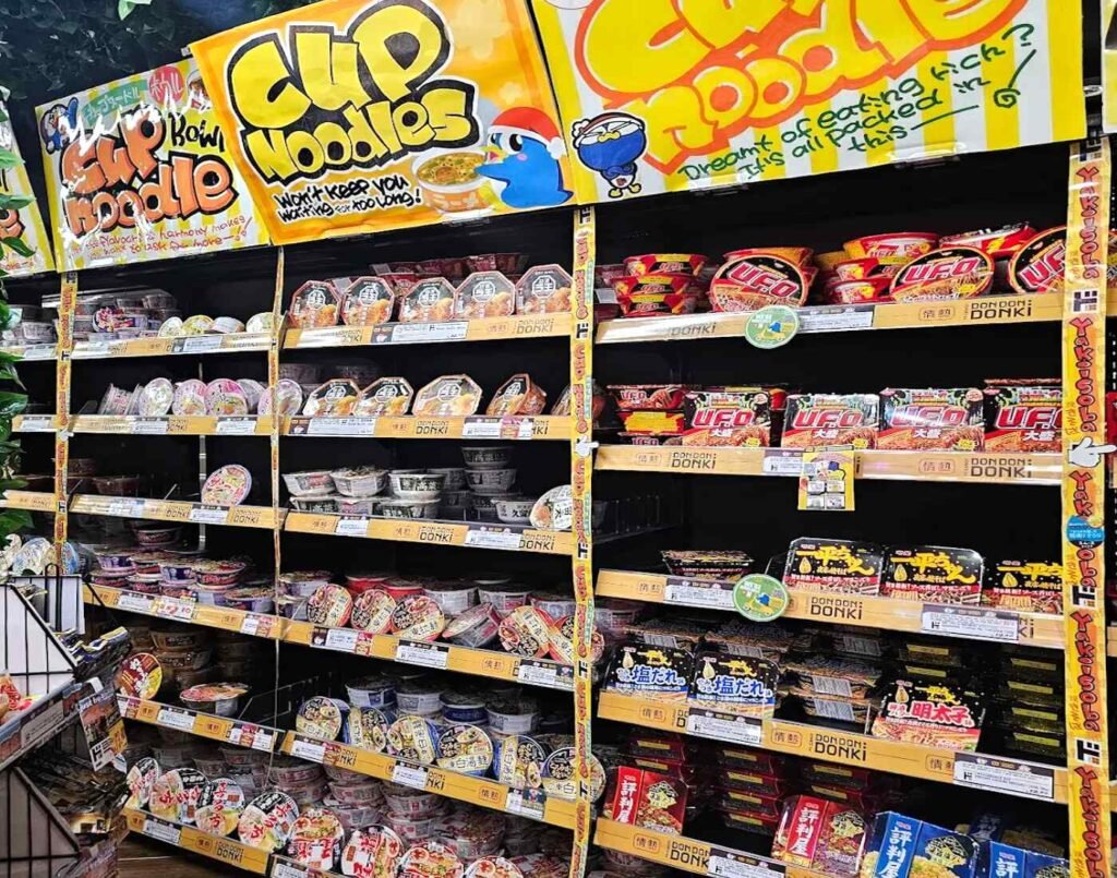 Shelves of Japanese cup noodles and yakisoba featuring Nissin Cup Noodle (Original, Seafood, Curry), U.F.O. Yakisoba and Donbei Udon, Jonetsu Kakaku Tonkotsu, Shoyu and Miso ramen bowls, Myojo Ippeichan and Hyoubanya Yakisoba, Acecook Super Cup 1.5x and Gotsumori Sauce Yakisoba, and Sapporo Ichiban cup noodles, on display at Don Don Donki Lot 10 in Bukit Bintang, Kuala Lumpur.