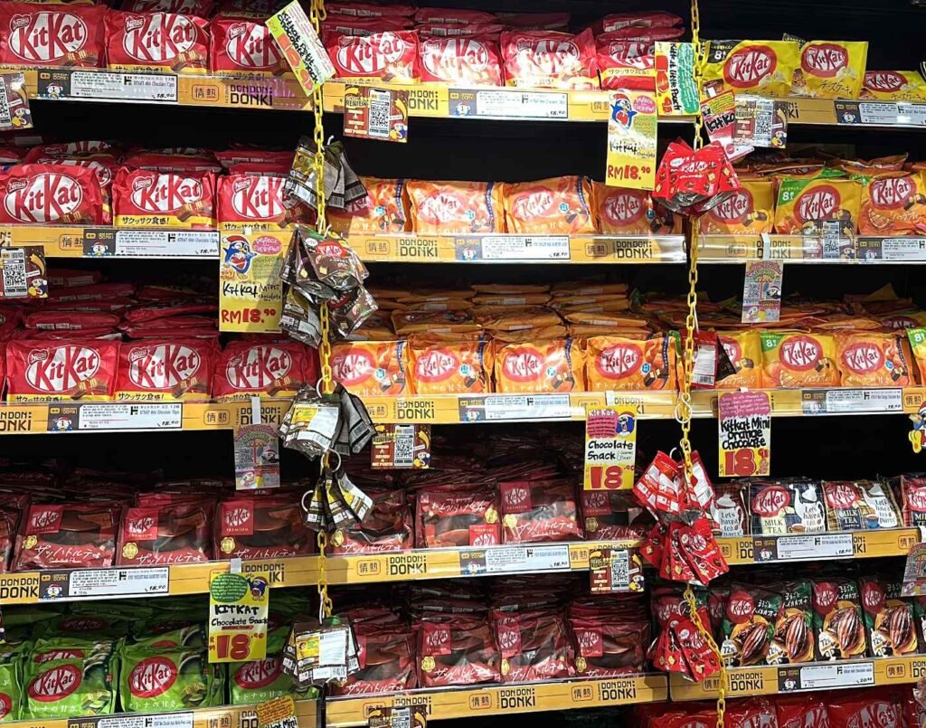 A colourful shelf display of assorted Japanese Kit Kat flavours including classic Milk Chocolate, Matcha Green Tea, Dark Chocolate, Caramel, and limited-edition varieties such as Sakura Strawberry and Cheesecake. The section features large family packs, mini boxes, and seasonal gift editions. Located on the Ground Floor of Jonetz By Don Don Donki Lot 10, a Japanese supermarket in Bukit Bintang, Kuala Lumpur.