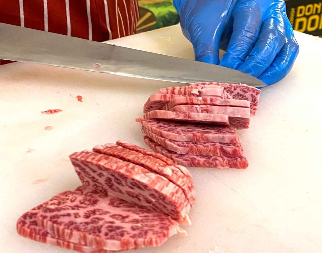 A butcher's gloved hands using a specialized knife to custom-slice a thick cut of raw Wagyu at the in-house meat cutting station, offering custom cuts for yakiniku grilling, shabu-shabu, and steak at Jonetz By Don Don Donki Lot 10, a Japanese supermarket in Bukit Bintang, Kuala Lumpur.