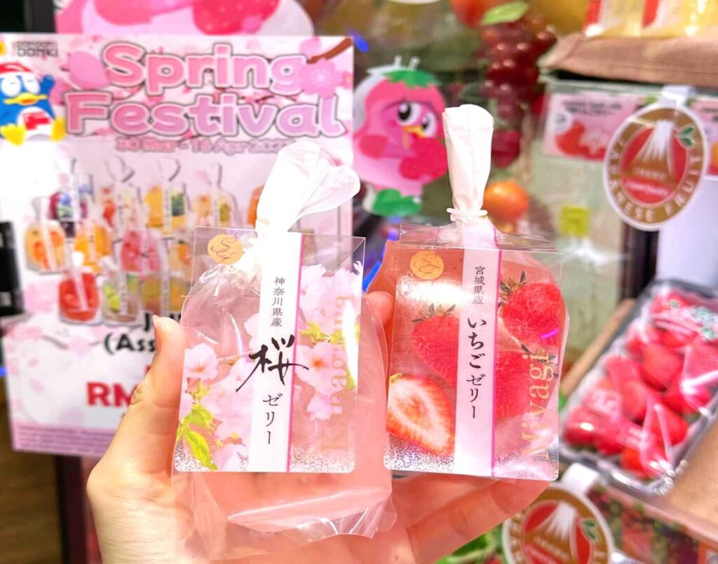 Shopper holding Furu Furu fruit jelly packs in Miyagi Strawberry and Kanagawa Sakura flavours in front of a Spring Festival promotion display at Don Don Donki Lot 10, Bukit Bintang.