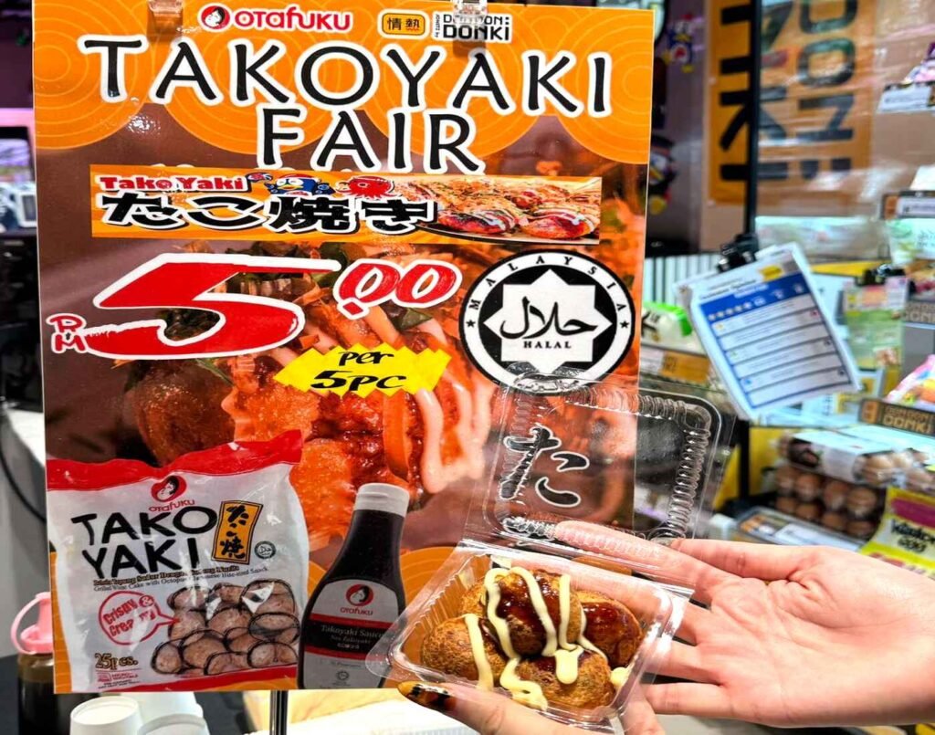 Freshly grilled takoyaki topped with Otafuku sauce held by a shopper in front of a Takoyaki Fair promotion sign showing RM5 for 5 pieces and a Halal logo at Don Don Donki Lot 10, Bukit Bintang.