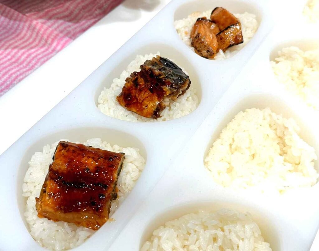 A chef uses white silicone molds for portioning sticky rice with glazed pieces of grilled Unagi (Eel) placed on top, a step in preparing gourmet rice balls or onigiri. A crowd favourite at Tomita Seimai, an Onigiri specialty restaurant located on the 2nd floor of Jonetz By Don Don Donki Lot 10, a Japanese supermarket in Bukit Bintang, Kuala Lumpur.