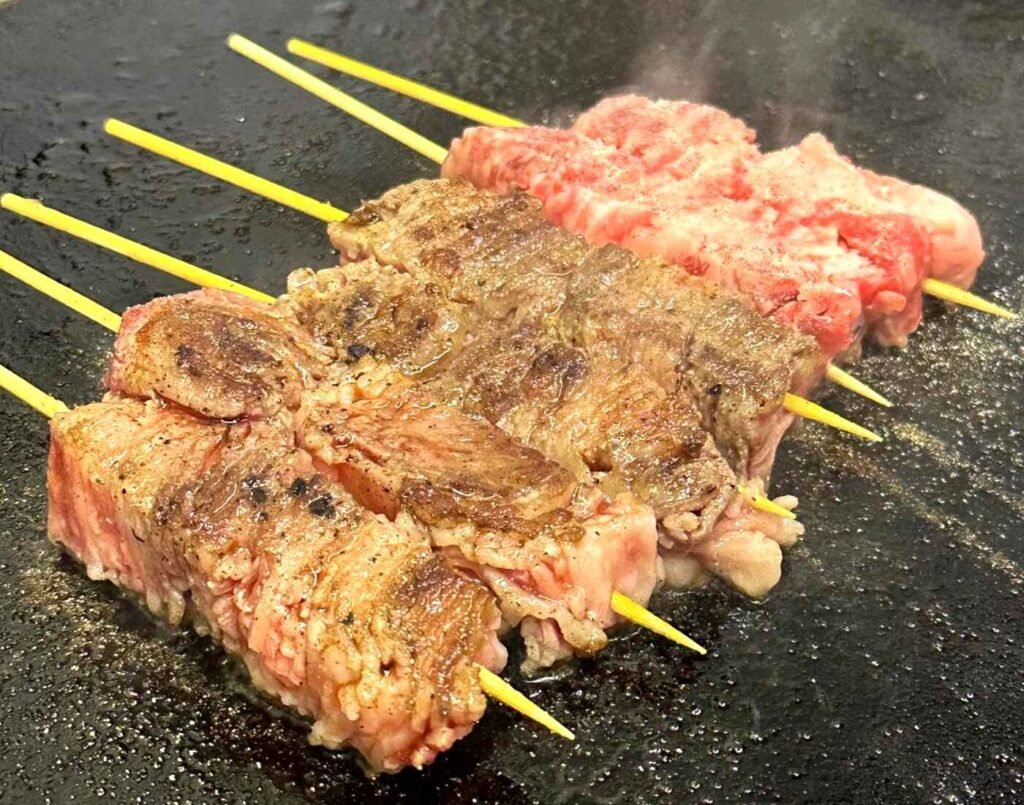 Thick wagyu beef skewers cooking and sizzling on a hot, black flat-top grill at the grilling station serving freshly grilled wagyu skewers, satay, and other ready-to-eat premium wagyu dishes at Jonetz By Don Don Donki Lot 10, a Japanese supermarket in Bukit Bintang, Kuala Lumpur.