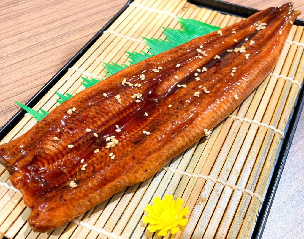 A close-up of premium Japanese grilled unagi (eel kabayaki) with tare sauce and sesame seeds on a bamboo mat, a seasonal offering at JONETZ by Don Don Donki Lot 10 for Doyo no Ushi no Hi (Day of the Ox).