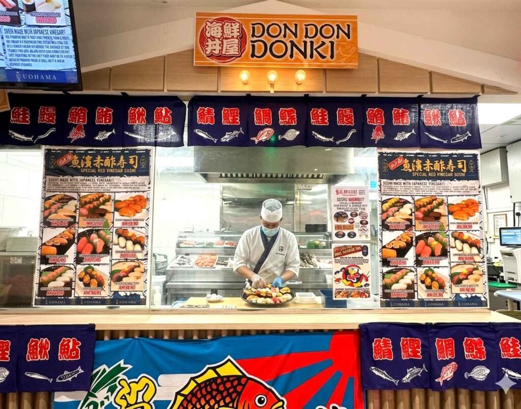 The main counter of Uohama Live Kitchen at JONETZ by Don Don Donki Lot 10, showing the red vinegar sushi menus, a la carte prices, and a chef working inside the kitchen.